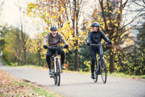 An active senior couple with electrobikes cycling outdoors on a road in park in autumn