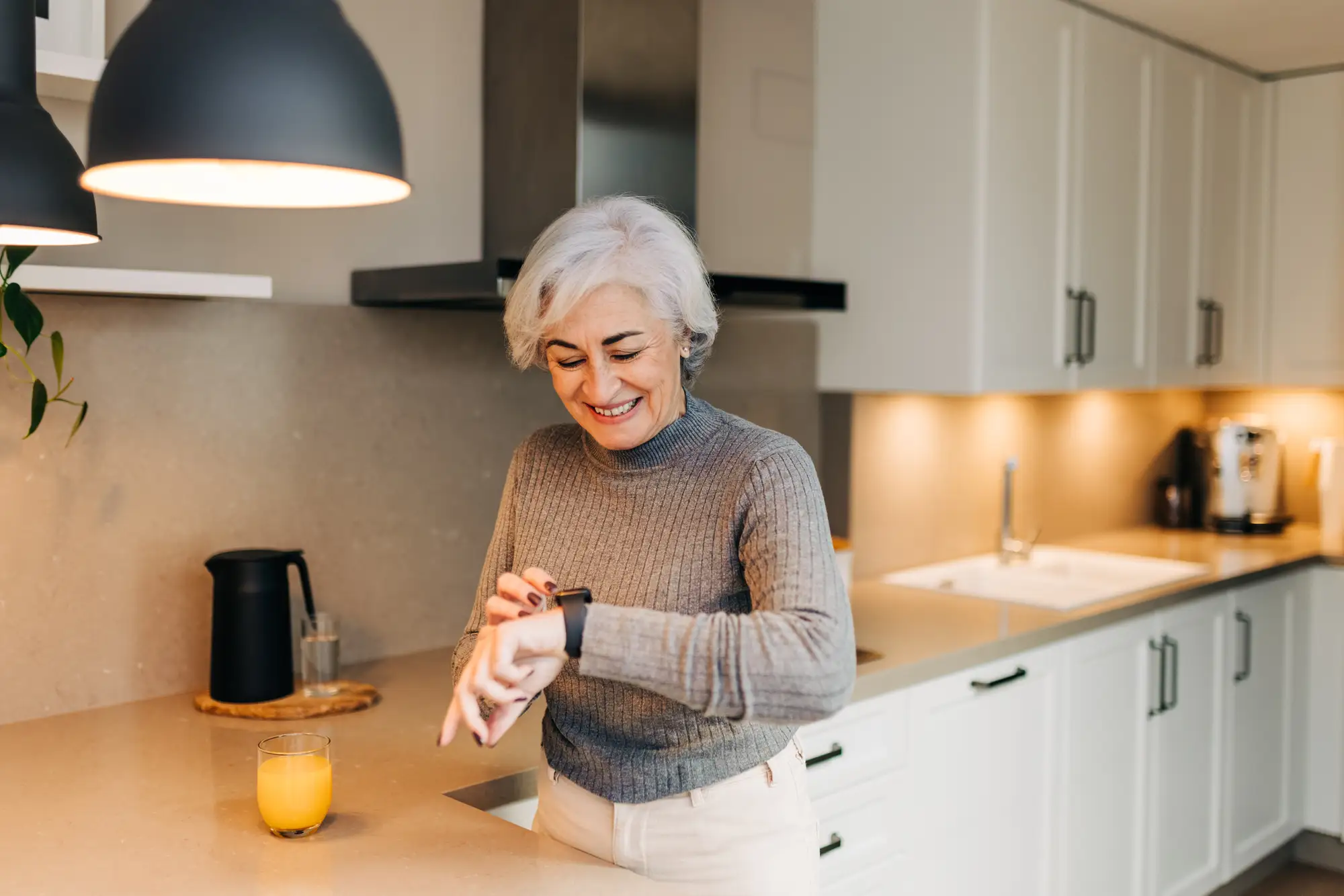 Healthy senior woman smiling at her smartwatch while standing in her kitchen. 