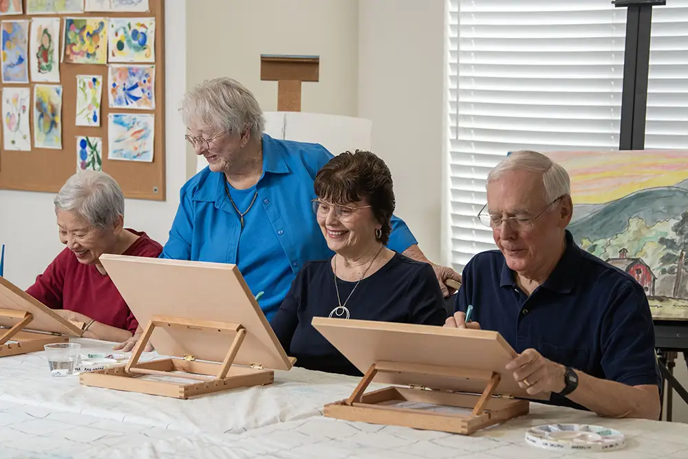Freedom Village residents sit together at a table during an art class in a vibrant hobby room, painting on tabletop easels while a finished watercolor landscape rests on a large easel behind them.