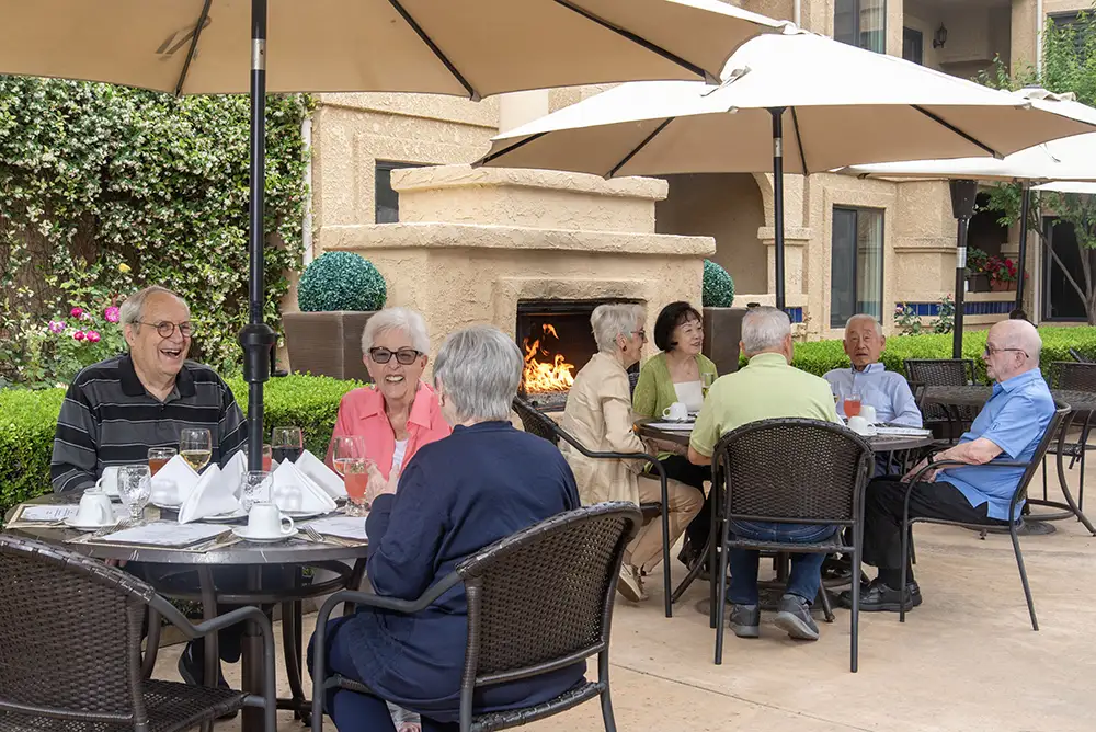 Seniors at Freedom Village gather for a meal at outdoor patio tables shaded by large umbrellas next to a roaring fireplace, enjoying a social afternoon with friends outside the community hobby room.