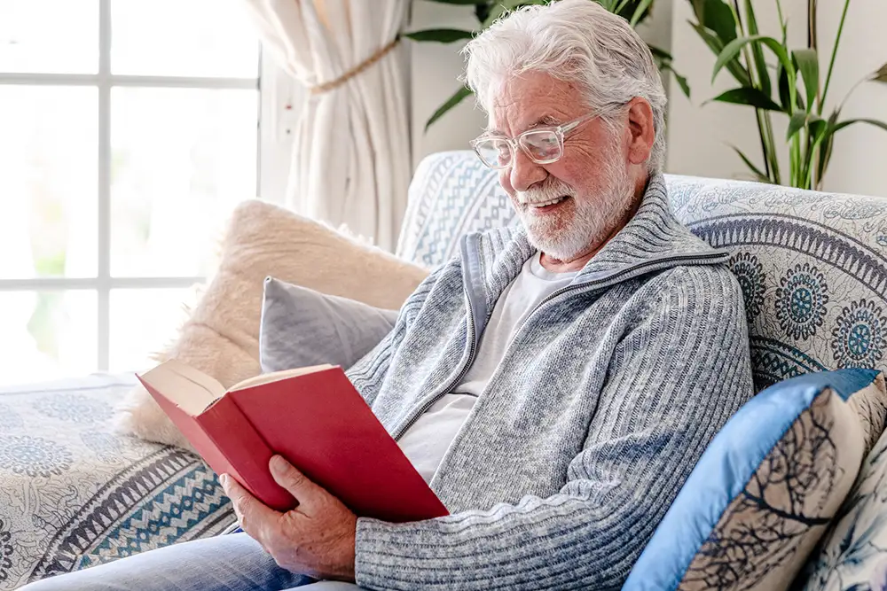 A senior at Freedom Village relaxes on a patterned couch with decorative pillows, smiling as he reads a hardcover book in a bright, sunlit community hobby room.